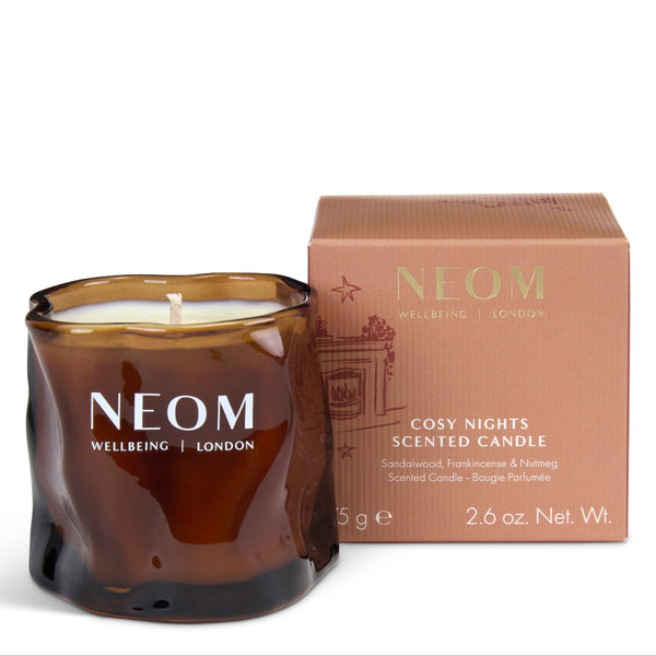 NEOM Cosy Nights scented candle in a dark amber glass jar next to its dusty pink packaging box, on a clean white background.