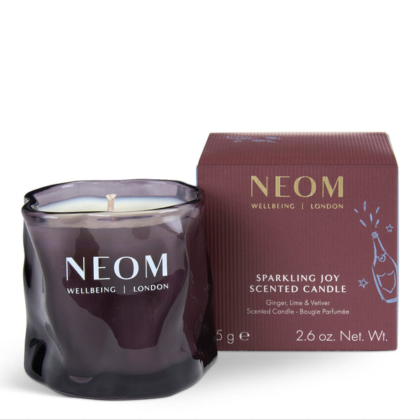 A NEOM Wellbeing London Sparkling Joy scented candle in a dark purple glass jar, shown next to its matching dark burgundy box on a clean white background.