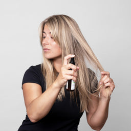 Woman applying hair product to her blonde hair against a plain background