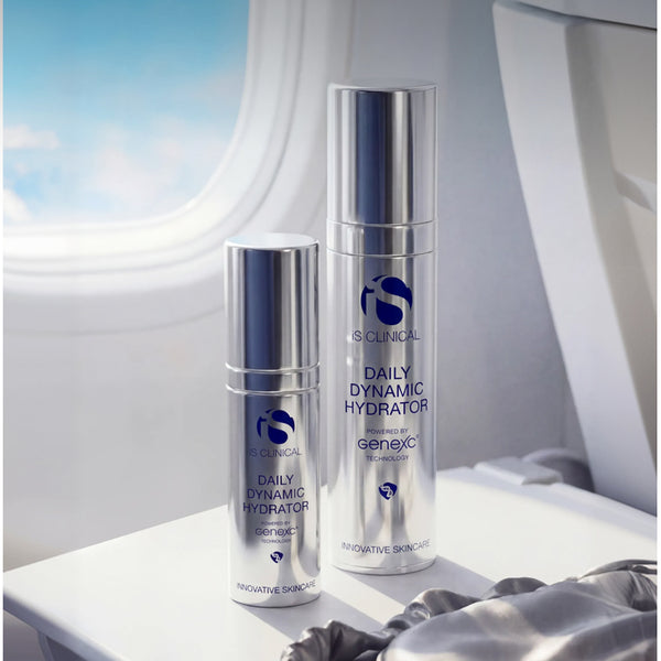 Two silver skincare bottles from IS Clinical Daily Dynamic Hydrator, one smaller bottle and one larger bottle, displayed on a white tray next to an airplane window.