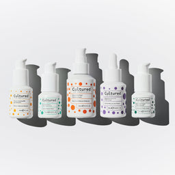 Five white pump bottles of Cultured Cosmetics skincare products arranged vertically in a staggered row against a light gray background with dark gray shadows.