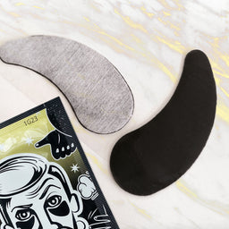 Two eye patches, one gray and one black, on a white surface with a product packaging featuring a face illustration.