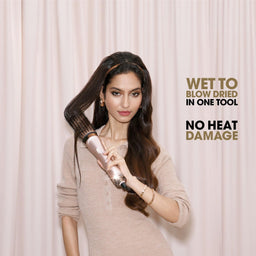 A woman with long dark hair holds a hair styling tool against her hair in front of a light pink curtain backdrop. Text on the right reads 'WET TO BLOW DRIED IN ONE TOOL' and 'NO HEAT DAMAGE'.