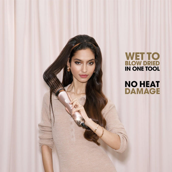 A woman with long dark hair holds a hair styling tool against her hair in front of a light pink curtain backdrop. Text on the right reads 'WET TO BLOW DRIED IN ONE TOOL' and 'NO HEAT DAMAGE'.