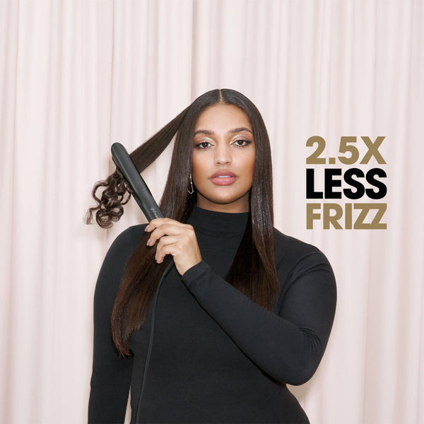 A woman with long dark hair wearing a black turtleneck holds a black hair straightener near a section of her hair, standing in front of a light pink curtain backdrop. Gold and black text on the right reads '2.