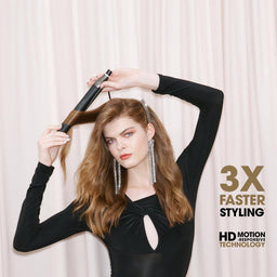 A woman with long wavy light brown hair styling her hair with a black hair straightener, wearing a black long-sleeve top against a cream-colored curtain backdrop. Text overlays on the right read '3X FASTER STYLING' and 'HD MOTION TECHNOLOGY'.