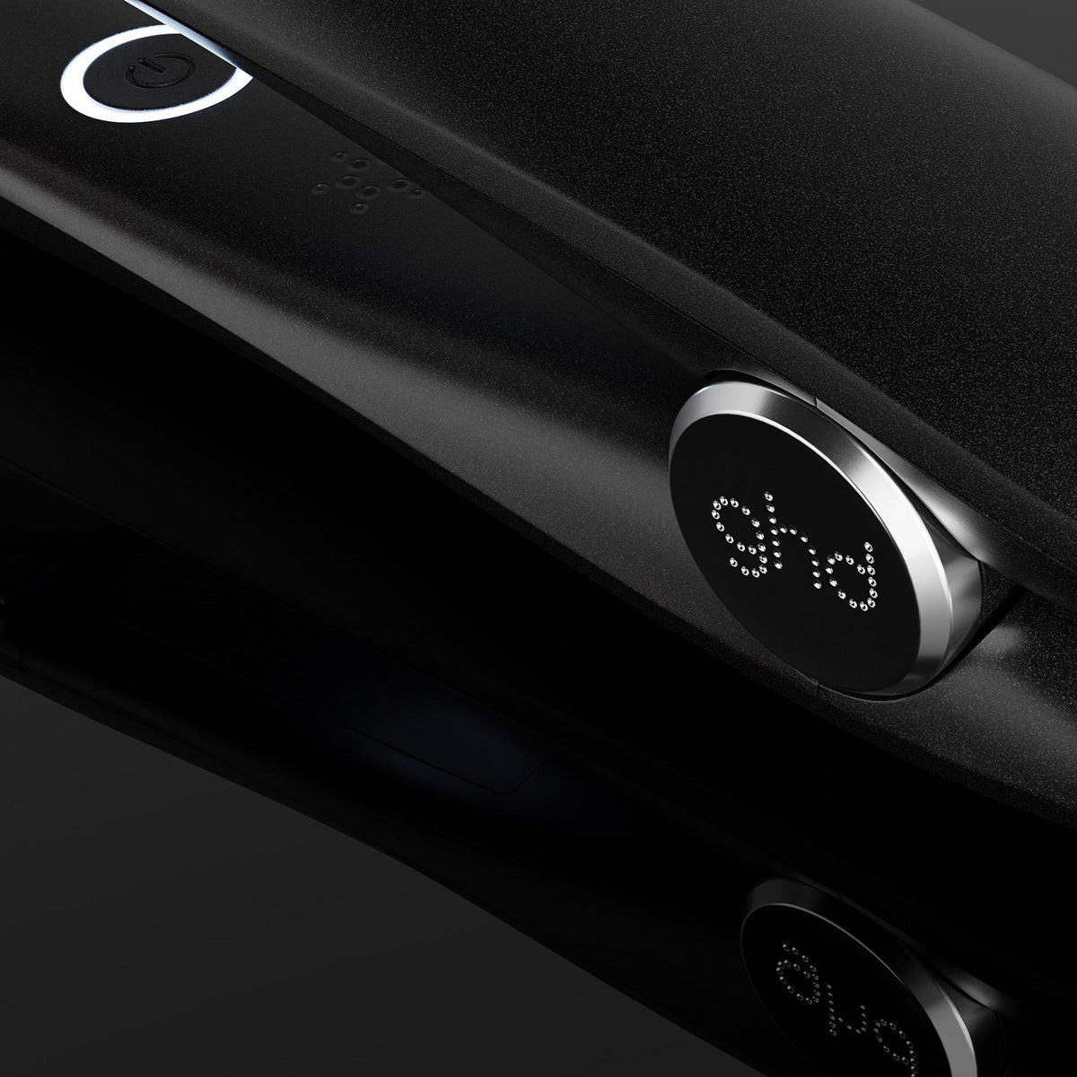 A close-up of a black ghd hair straightener with its power cord, showing the brand logo on the control dial against a dark reflective background.