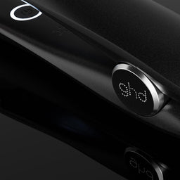 A close-up of a black ghd hair straightener with its power cord, showing the brand logo on the control dial against a dark reflective background.