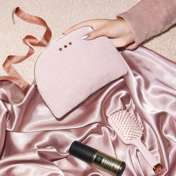 A hand holds a blush pink makeup pouch with three metal eyelets, styled on draped satin fabric with a pink hairbrush and a black-and-gold cosmetic tube nearby.