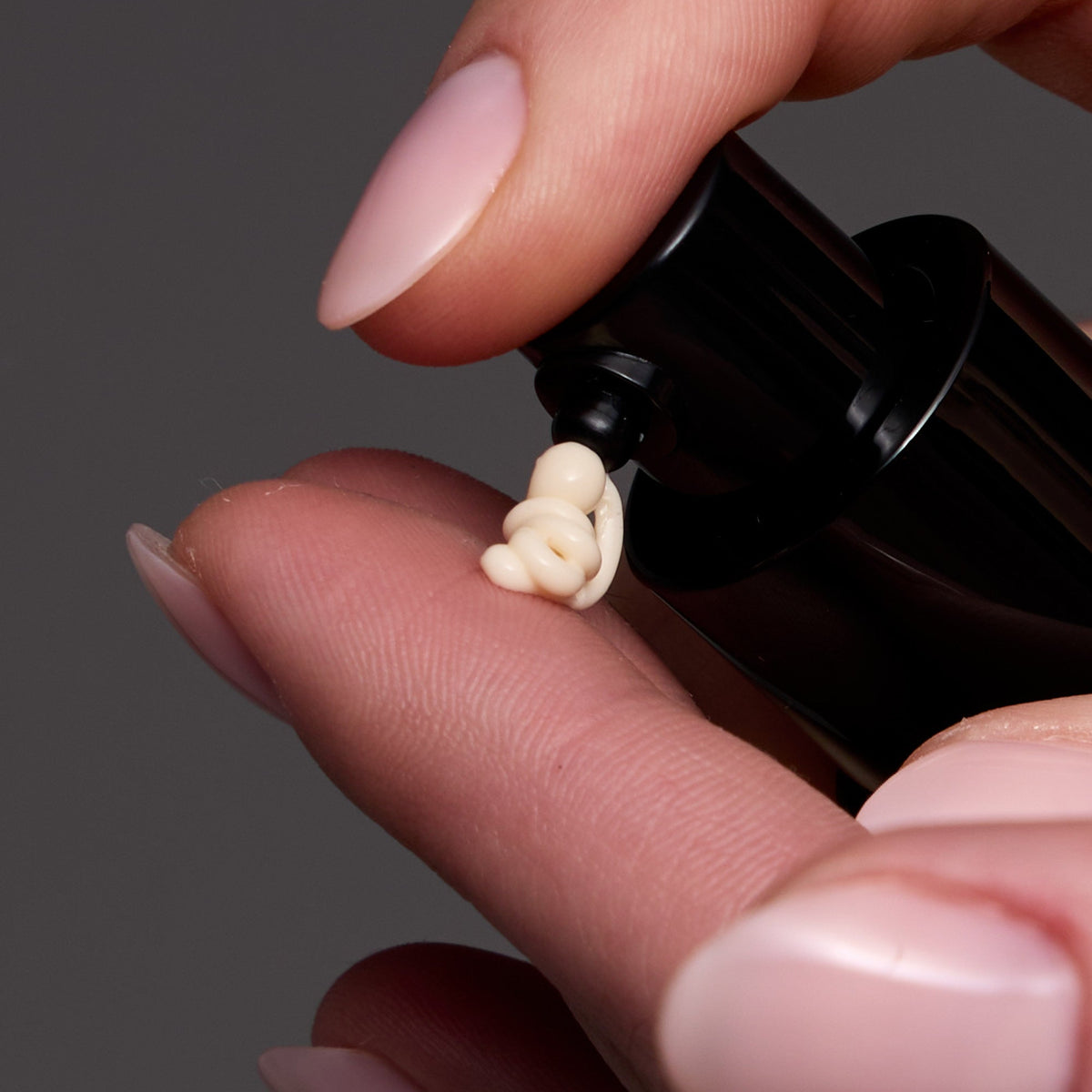 A close-up of a hand with pale pink manicured nails dispensing a small dollop of cream-colored cosmetic product from a black pump bottle against a dark gray background.