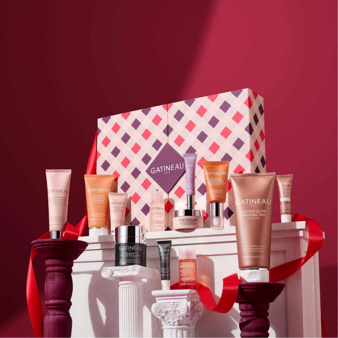 A Gatineau skincare gift set arranged on white pedestals against a deep burgundy background. The set includes multiple skincare tubes, bottles, and a patterned gift box with a red ribbon, featuring brands like Gatineau and L'Oréal Paris.
