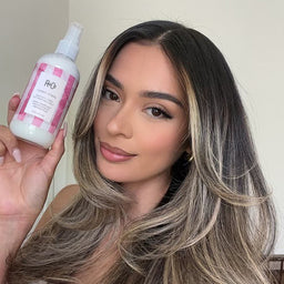 Woman with long, wavy hair holding a bottle of R+Co hair product.