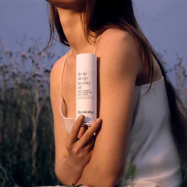 A woman outdoors at sunset holds a white cylindrical bottle labeled 'deep sleep beauty oil' against her shoulder. The bottle is branded 'thisworks'.