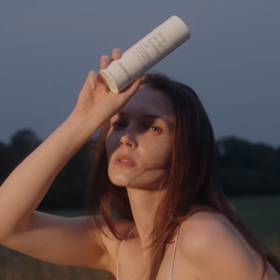A woman outdoors at dusk holds a white cylindrical bottle against her forehead. The bottle has black text that appears to read 'hair oil' and 'bionica'.