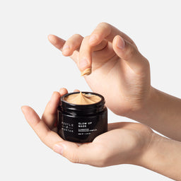 Hand holding a jar of 'Glow Up Mask' with a small amount on a light background