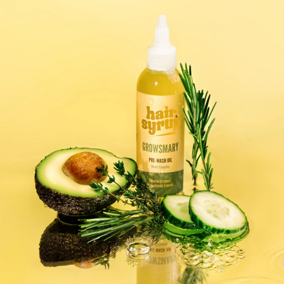 Bottle of 'hair syrup' Growsmary Pre-Wash Oil with an avocado, cucumber, and rosemary on a yellow background