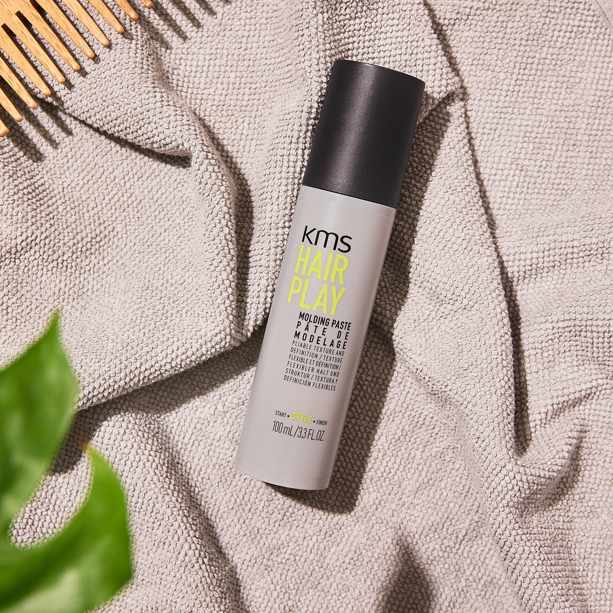 KMS Hair Play product on a textured surface with a comb and plant leaf.