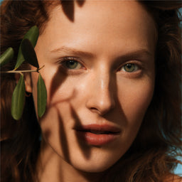 Close-up of a woman's face with a leaf in front of her eye, sunlit.