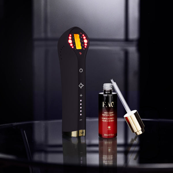 A black handheld beauty device with illuminated red and yellow indicator lights stands beside a small red cosmetic bottle and a gold-and-white applicator on a glossy black round table against a dark studio background.