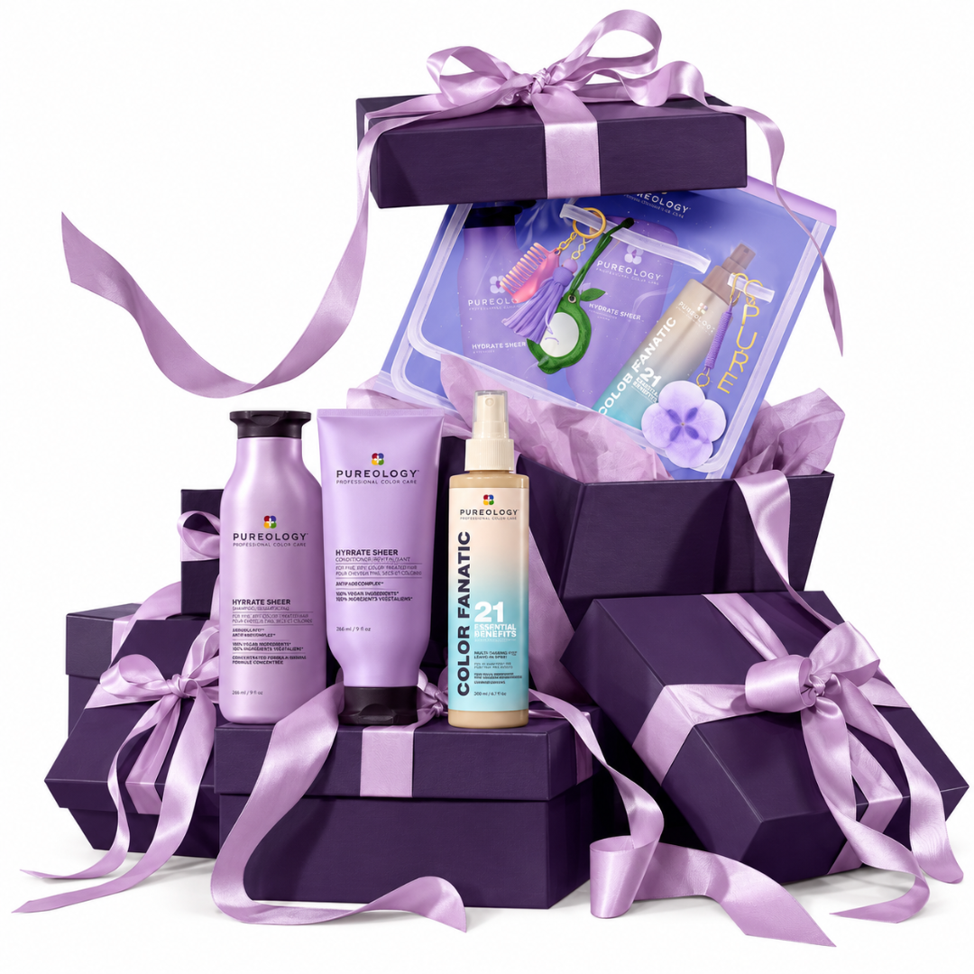 A haircare gift set arranged in stacked purple gift boxes with lavender satin ribbons on a white background. Visible products include Pureology Advanced Biotin Shampoo, Advanced Breaker, Color Fanatic 1-4 Color Protect Spray, and a purple hair color treatment kit booklet.