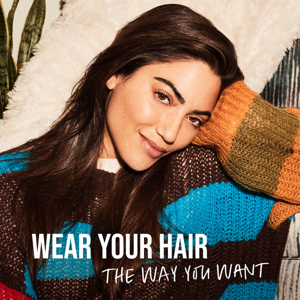 Woman with long hair leaning against a textured surface with text 'WEAR YOUR HAIR THE WAY YOU WANT'.