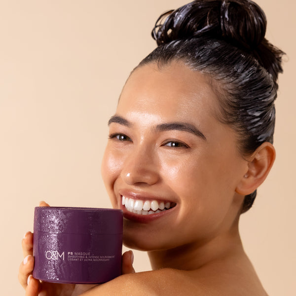 A smiling woman with dark hair in a bun holds a purple O&M Paris Masque jar next to her face against a warm beige background.