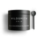 A matte black jar of Wildsmith Skin Dual Action Exfoliating Treatment, shown next to a matching black spoon on a clean white background.