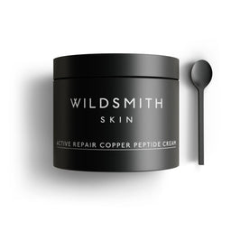 A matte black cosmetic jar labeled 'WILDSMITH SKIN' with white text reading 'ACTIVE REPAIR COPPER PEPTIDE CREAM,' shown on a white background with a matching black spoon to the right.