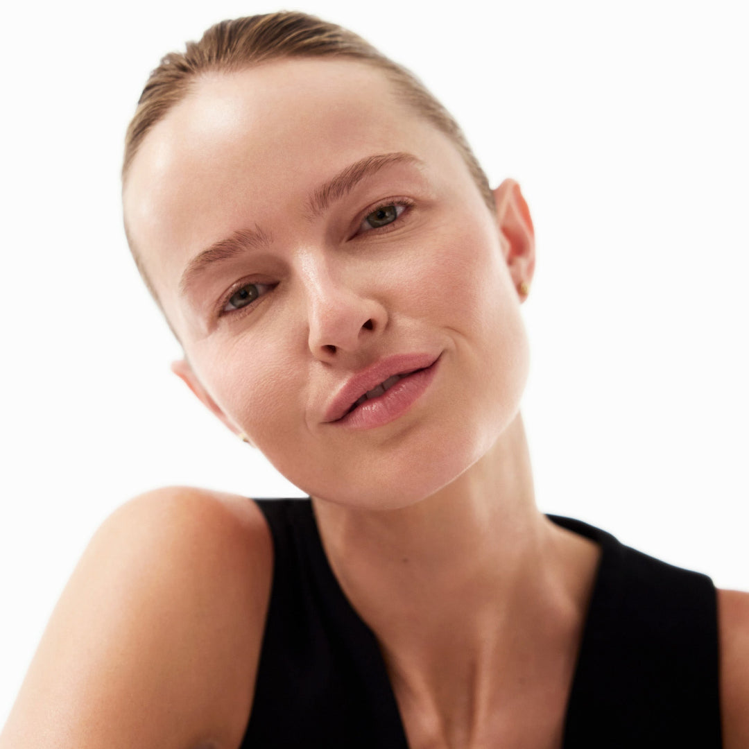 A close-up portrait of a woman with her hair pulled back, wearing a black sleeveless top against a plain white background.