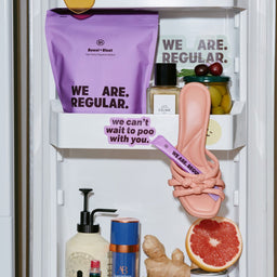 A styled refrigerator door scene with multiple props and text-based branding. The most salient sold item appears to be the pink slip-on shoe with a purple strap hanging on the middle shelf, surrounded by other objects including a purple Sweet Rusal bag, a perfume bottle, olives, ginger, a grapefruit slice, and Perello canned soda.