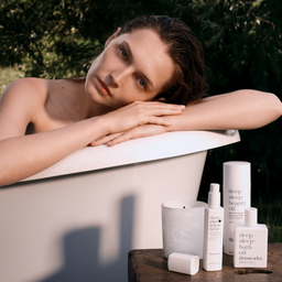 A woman rests her head on her arms across the edge of a white bathtub in an outdoor setting with green foliage in the background. Several white skincare and bath products are arranged on a wooden surface in the foreground, including bottles and a candle.