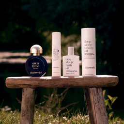 Four skincare products from thisworks are arranged on a rustic wooden stool outdoors against a blurred green garden background. The set includes a round dark blue bottle with a gold cap labeled 'own time', a white spray bottle, a white bath oil bottle, and a tall white beauty oil bottle.