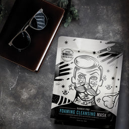 Foaming cleansing mask packaging with a stylized face design on a dark surface with sunglasses and a book.