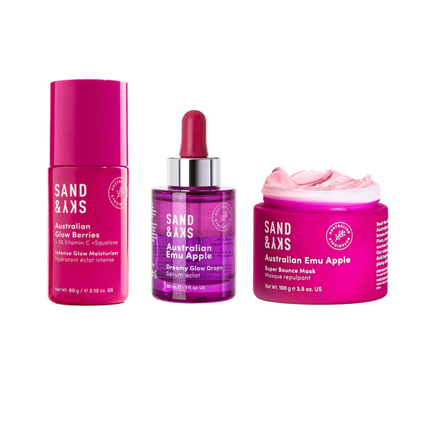 Three SAND & AXS skincare products arranged side by side on a plain white background: a pink Glow Berry serum bottle, a purple emulsion dropper bottle with a red dropper top, and a bright pink emu apple sheer bounce mask jar with the lid off and pink cream visible.