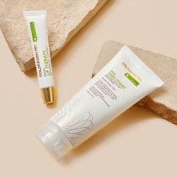 Two cosmetic tubes from Gold Skin Bio on a beige background with decorative stone props. One tube is a white lip therapy tube with a gold cap, and the other is a white hand cream tube with a clear cap.