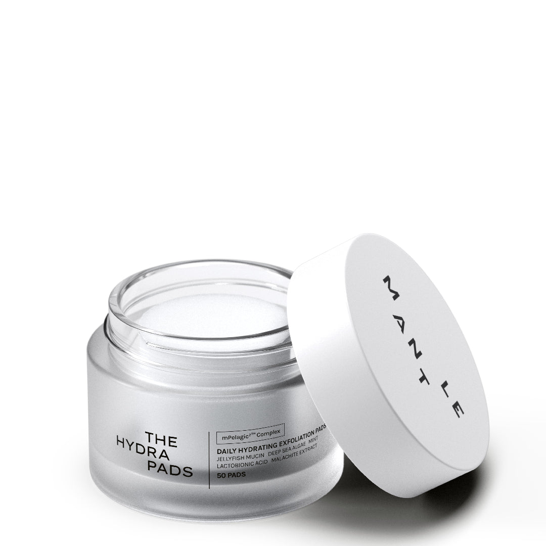 An open jar of MANTLE The Hydra Pads with its white lid leaning against it, showing the white cream inside. The jar is made of frosted clear glass, and the lid features the brand name 'MANTLE' in black text.