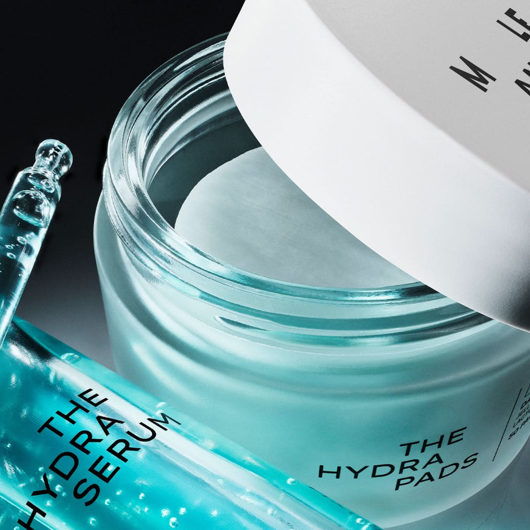A close-up of a clear cosmetic jar labeled 'THE HYDRA PADS' with a white lid partially off, alongside a blue glass dropper bottle labeled 'THE HYDRA SERUM' on a dark background.