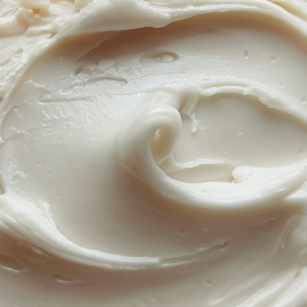 A close-up texture shot of a thick, creamy off-white cosmetic cream with smooth swirls and soft shadows.