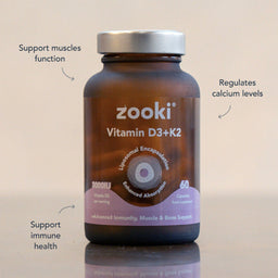 A brown supplement bottle labeled 'zooki Vitamin D3+K2' with a silver cap, shown on a light beige background with three callout text overlays describing benefits. The bottle is centered and the text points to muscle function, calcium levels, and immune health.