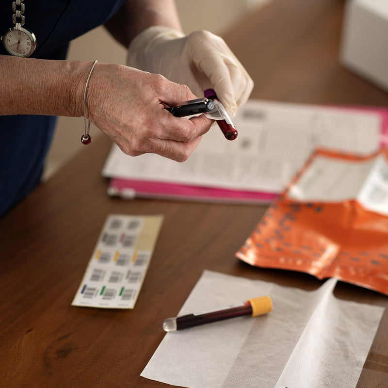 Person holding a blood sample with gloves on, surrounded by medical supplies on a wooden surface.