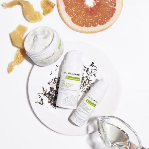 Three white skincare products from the brand GELMAGGINI are arranged on a white plate with grapefruit peel and a sliced grapefruit, alongside a jar of white cream. A hand holding a small glass pitcher is visible in the bottom right corner, set against a clean white background.